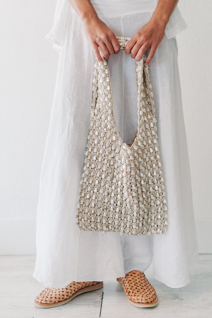 A person wearing a long white dress and woven tan slip-on shoes holds a large chunky knit shoulder bag in cream and beige.