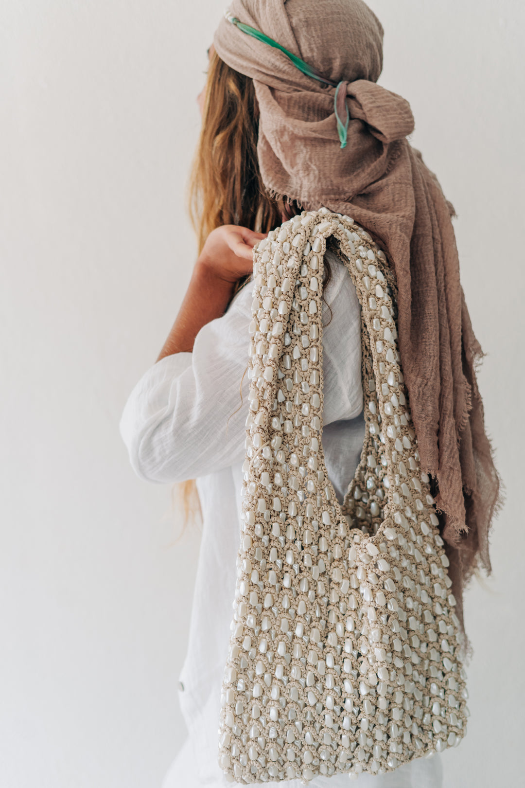 A woman wearing a brown headscarf and white top carries a large chunky knit shoulder bag in a light beige/cream color, shown from the back against a plain white background.