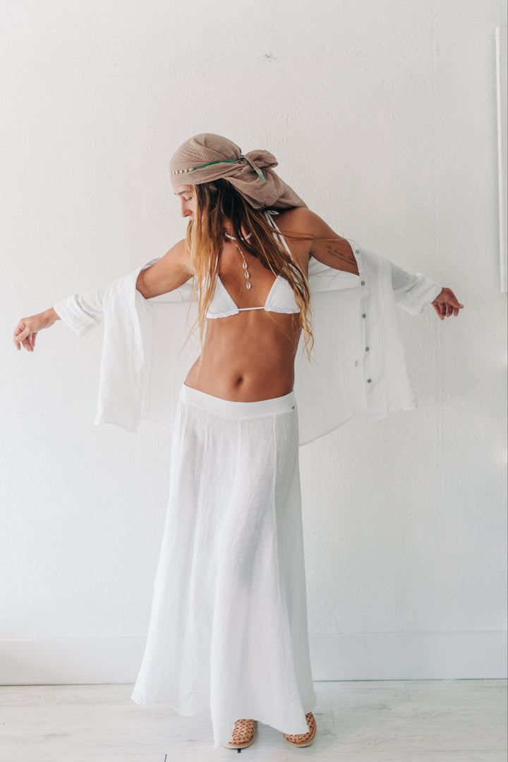 A woman stands with her back to the camera in a bright white room, wearing a white bikini, a long white maxi skirt, a white open-front cardigan, and a beige headscarf.