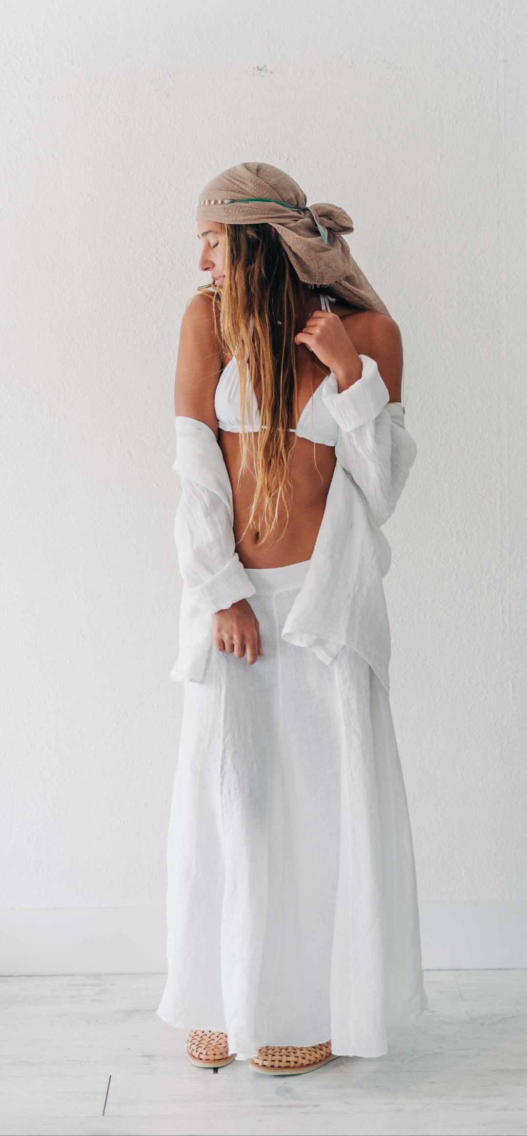 A woman stands with her back to the camera wearing a white two-piece outfit consisting of a bikini-style top and a long flowing skirt, with a beige headscarf tied around her hair and beige woven sandals.