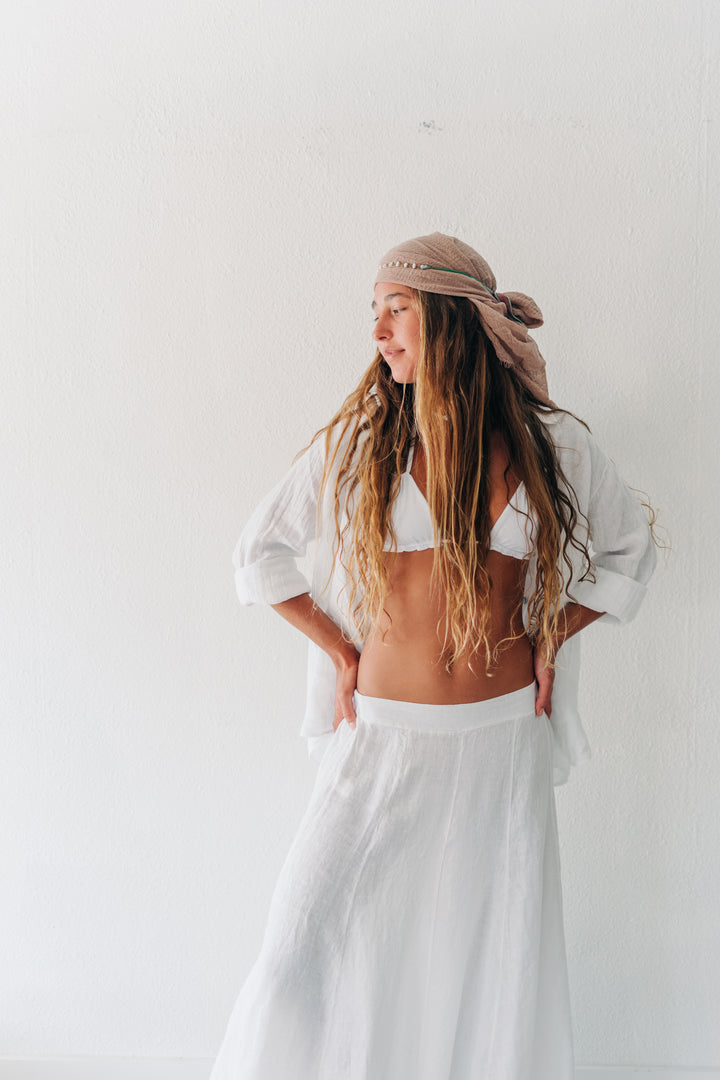 A woman with long wavy light brown hair wearing a white bikini, a loose white shirt, a long white skirt, and a beige patterned headscarf, posing against a plain white wall.