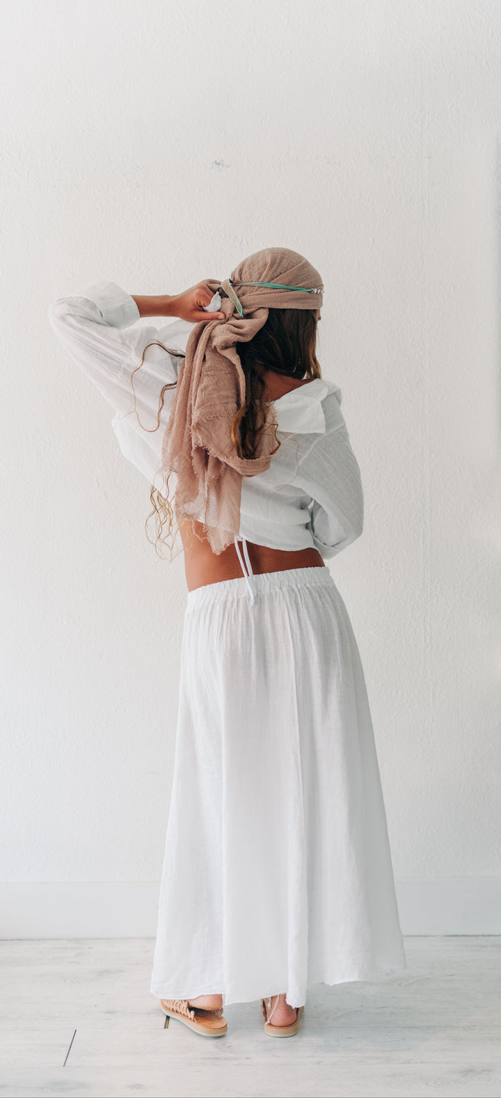 A woman stands with her back to the camera in a bright white room, wearing a white long-sleeve crop top and a long white skirt. She is adjusting a beige headscarf with a matching beige bag slung over her shoulder.