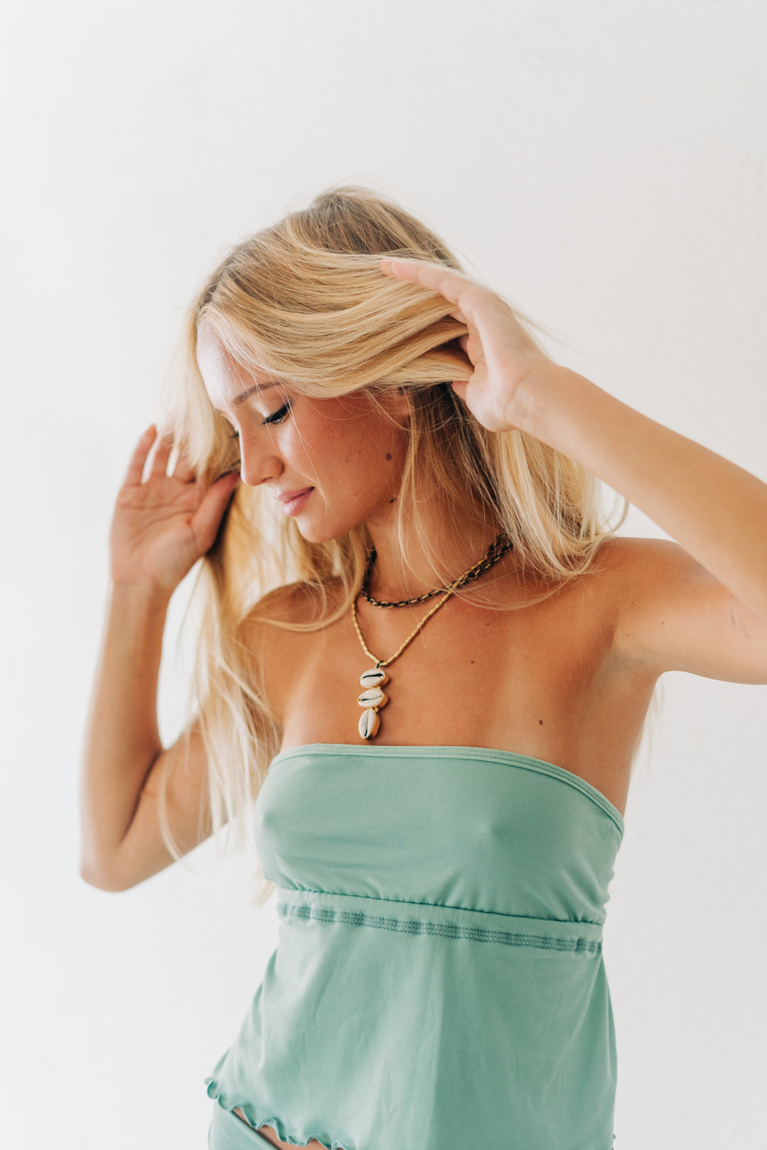 A blonde woman wearing a mint green strapless top and layered necklaces, posing against a plain white background.