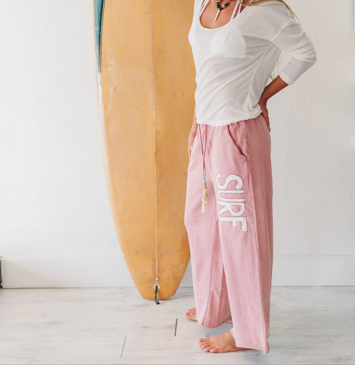 A woman stands barefoot in a white room wearing a green beanie, white long-sleeve top, and pink wide-leg sweatpants with &