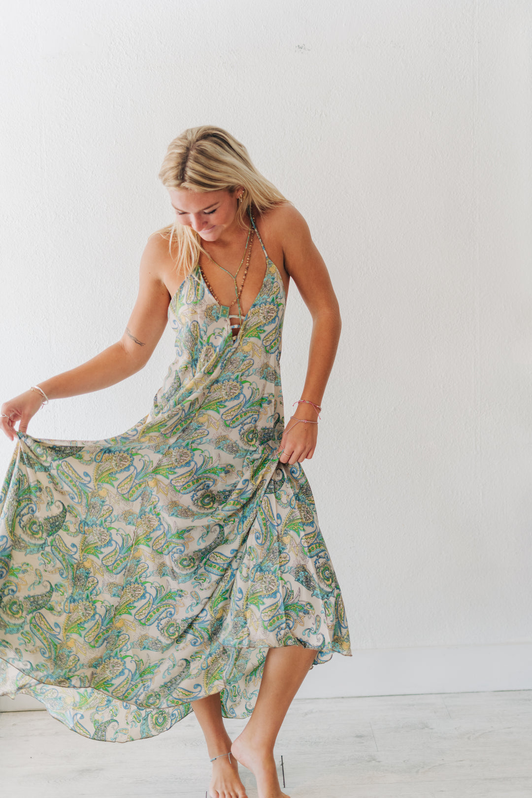 A woman wearing a sleeveless midi dress with a green, blue, and beige paisley pattern, holding out the skirt to show its flow against a plain white wall.