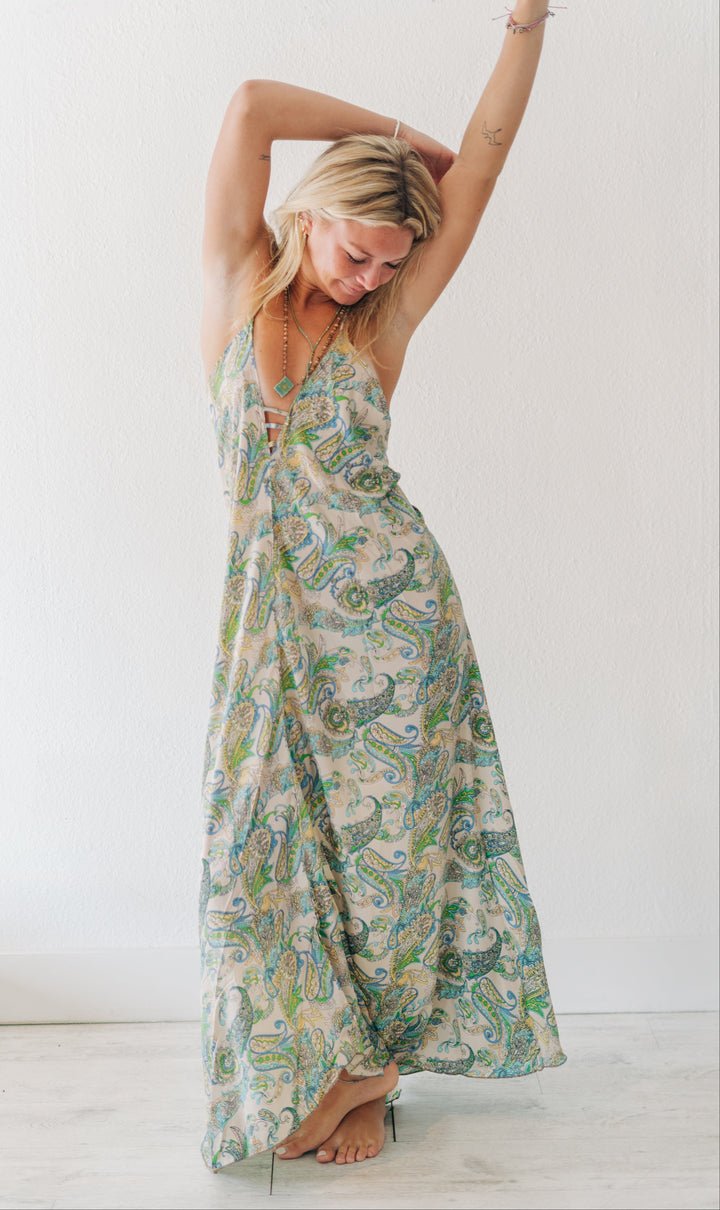A woman modeling a sleeveless maxi dress with a green, blue, and beige paisley pattern, posing with her arms raised against a white wall.