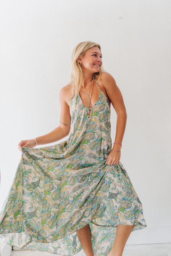 A woman modeling a sleeveless, V-neck maxi dress with a green, blue, and beige paisley pattern, holding out the skirt to show its flow against a plain white wall.