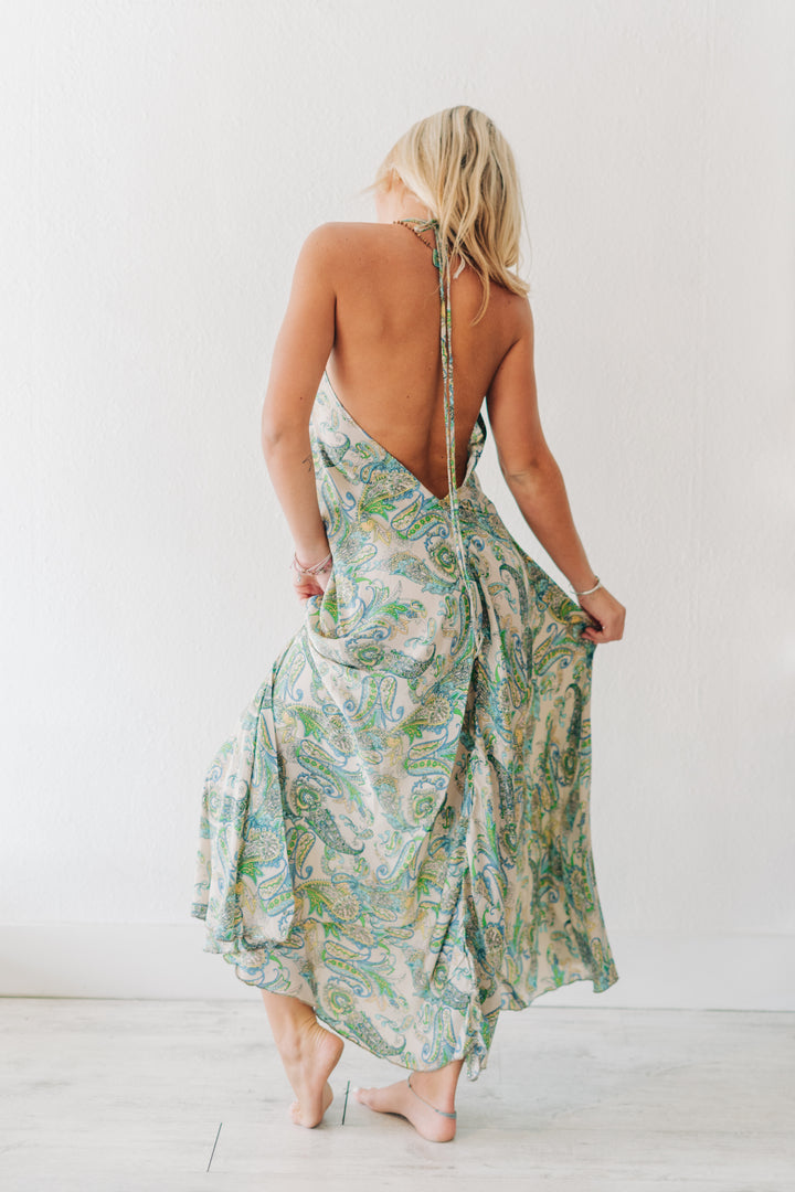 A woman shown from behind wearing a sleeveless, open-back maxi dress with a green and blue paisley pattern on a cream background, standing barefoot against a white wall.