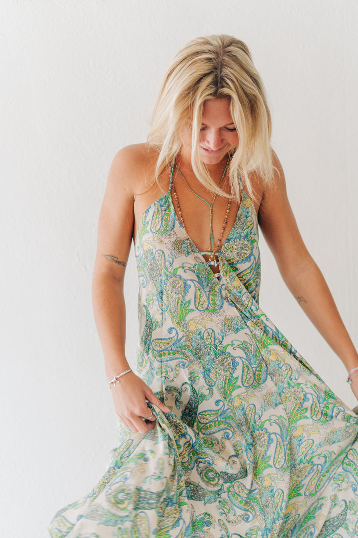 A woman wearing a sleeveless maxi dress with a green and blue paisley pattern on a cream background, standing against a plain white wall.