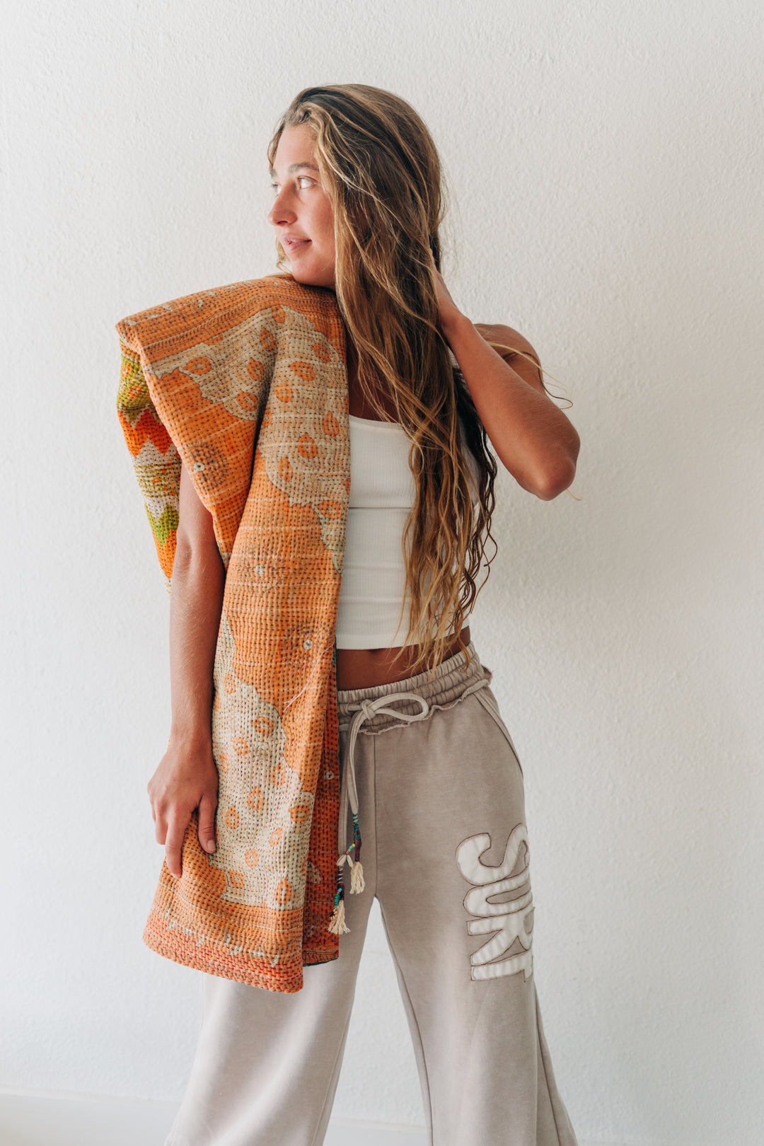 A woman stands against a white wall wearing beige drawstring sweatpants with white &