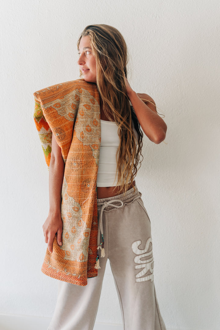 A woman stands against a white wall wearing beige drawstring sweatpants with white &