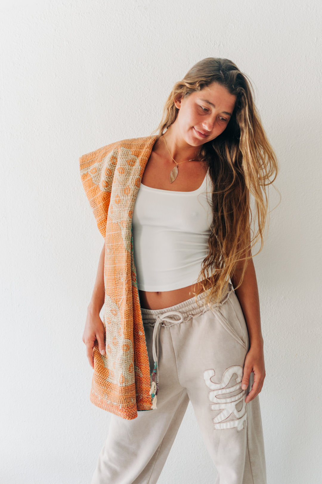 A woman wearing a white ribbed tank top and light beige sweatpants with a white graphic print, draped with an orange and beige patterned knit blanket against a white wall.