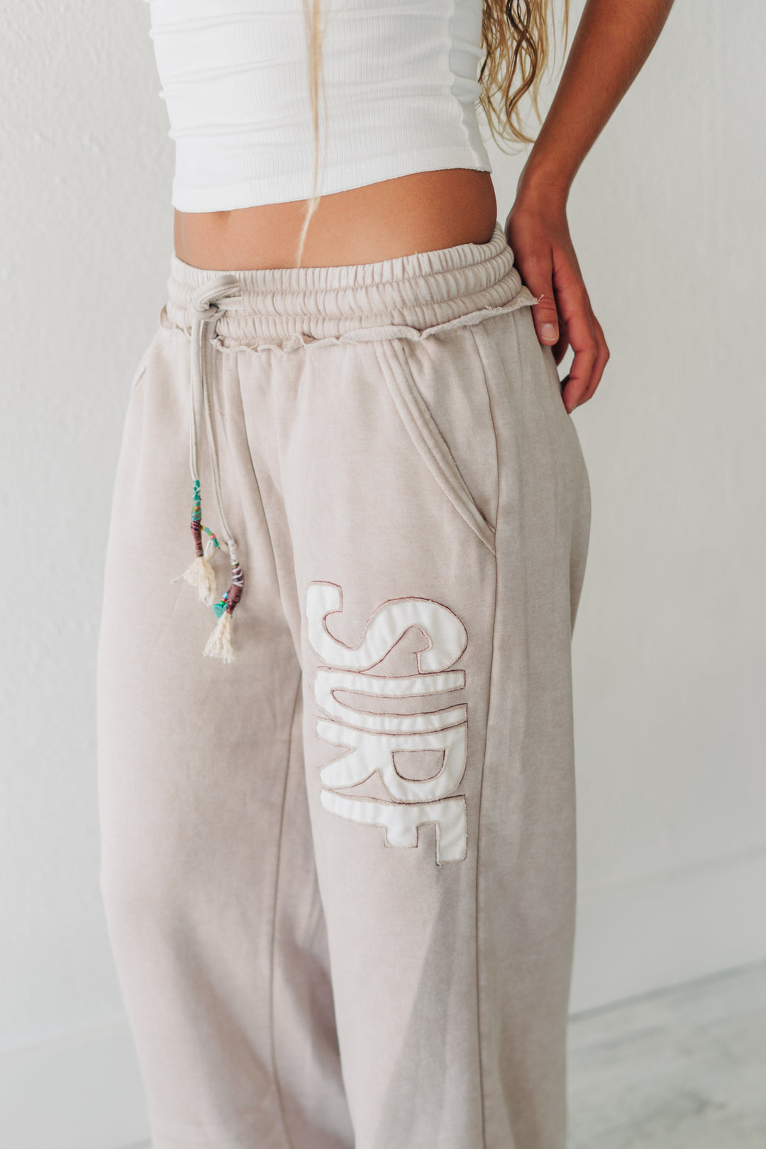 A person wearing light beige lounge sweatpants with a large white embroidered &