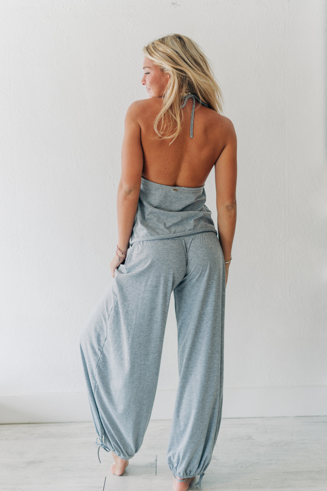 A woman shown from behind wearing a light gray heathered two-piece lounge set consisting of a halter-style top and matching wide-leg pants, standing against a plain white wall.