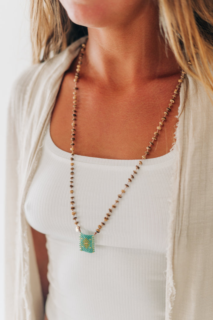 A close-up of a woman wearing a long beaded necklace with a turquoise and gold square pendant over a white ribbed tank top and a light beige cardigan.