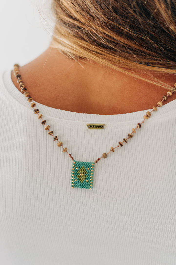 A close-up lifestyle shot of a model from the back wearing a beaded necklace. The necklace features a rectangular turquoise and gold beaded pendant, accented by small brown and gold beads.