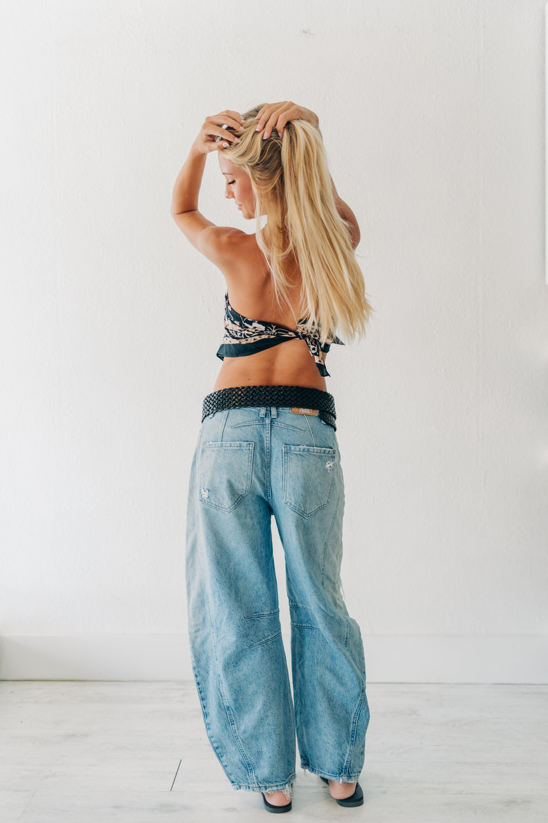 A woman with long blonde hair is shown from behind wearing light-wash wide-leg denim jeans, a black and white patterned halter top, and a black belt, standing against a plain white wall.