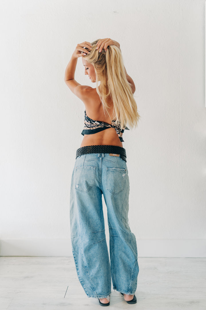 A woman with long blonde hair is shown from behind wearing light-wash wide-leg denim jeans, a black and white patterned halter top, and a black belt, standing against a plain white wall.