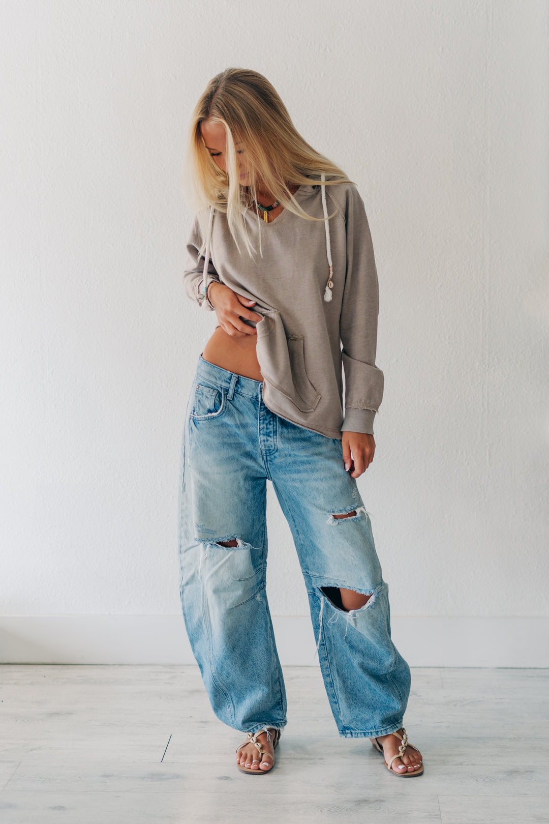 A woman modeling a taupe gray oversized hoodie and distressed light blue wide-leg jeans with ripped knees, paired with brown sandals, standing against a white wall.