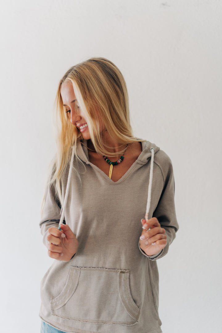 A woman wearing a light taupe V-neck pullover hoodie with white drawstrings and a front kangaroo pocket, standing against a plain white wall.
