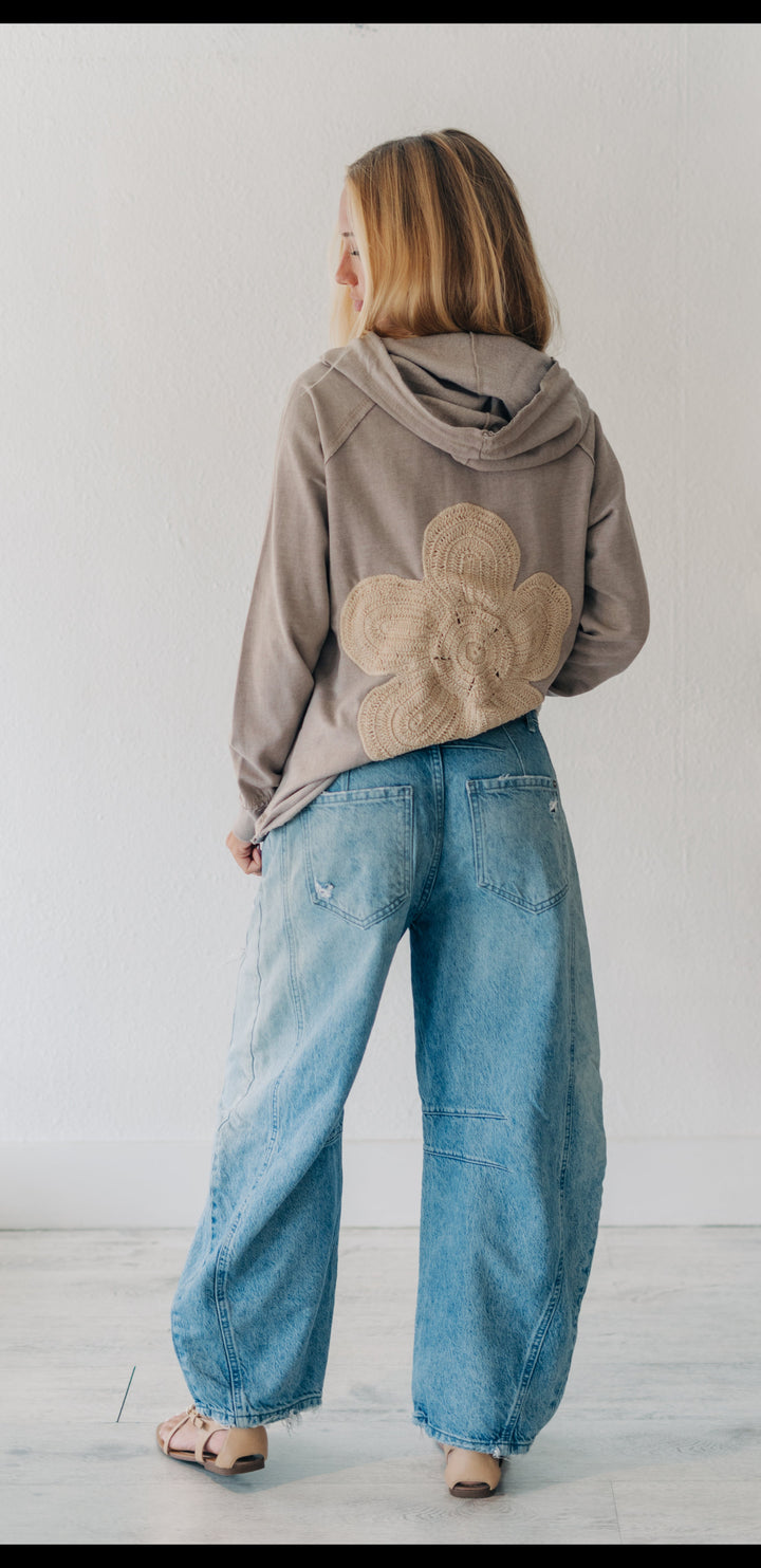 A woman viewed from behind wearing a taupe-brown hooded sweatshirt with a large beige flower applique on the back, paired with wide-leg light blue denim jeans and flat sandals, standing in a bright indoor room.