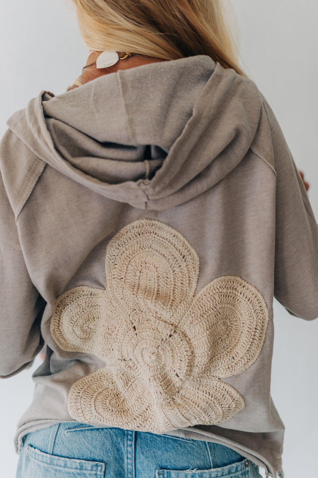 A woman viewed from behind wearing a taupe-gray hooded sweatshirt with a large beige crocheted floral applique on the back, paired with blue jeans.