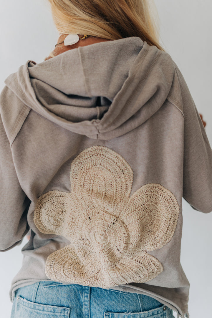 A woman viewed from behind wearing a taupe-gray hooded sweatshirt with a large beige crocheted floral applique on the back, paired with blue jeans.