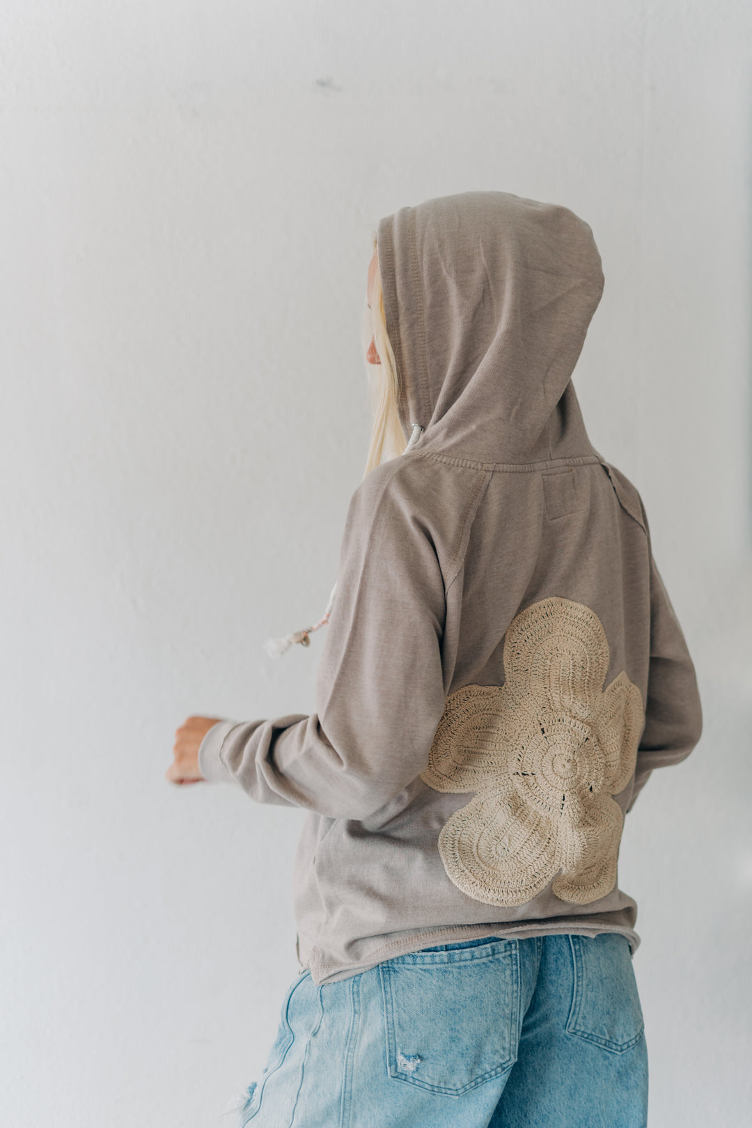 A person shown from the back wearing a taupe-brown hooded sweatshirt with a large beige woven flower applique on the back, paired with light blue jeans, against a plain white wall.