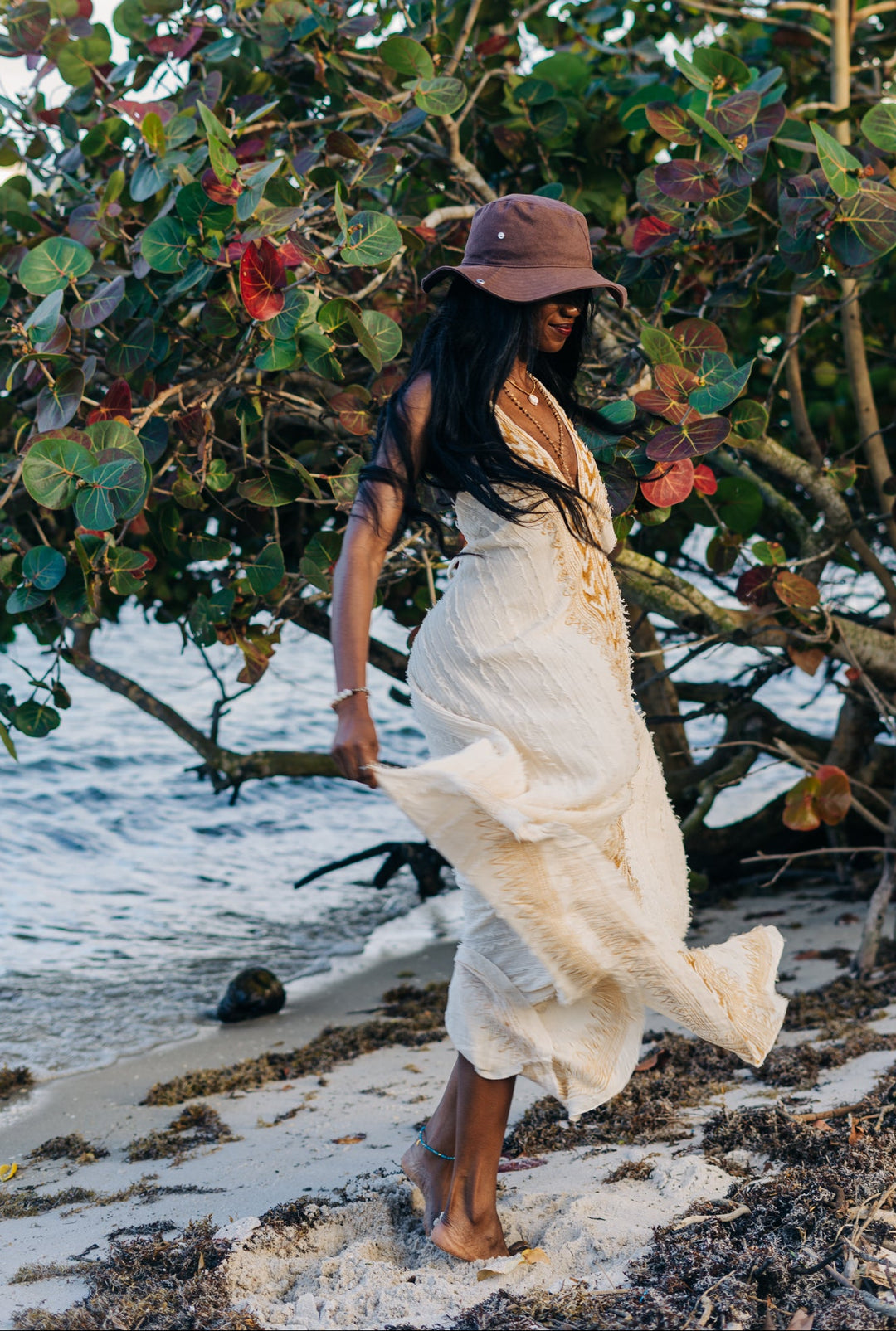 Woman in a white dress and brown hat standing on a beach with trees and water in the background