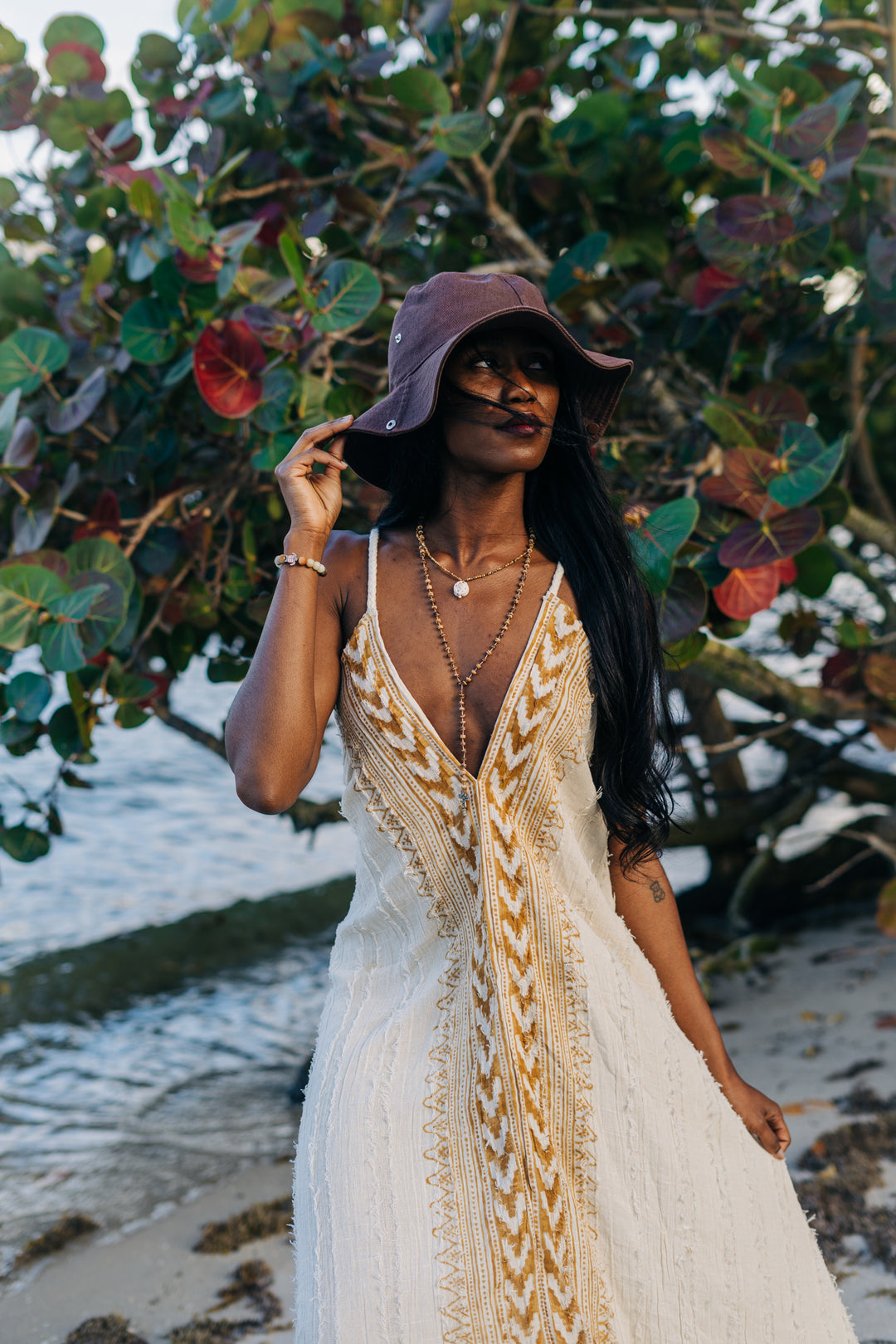 Woman in a white dress with gold patterns and a purple hat standing in front of a leafy background.