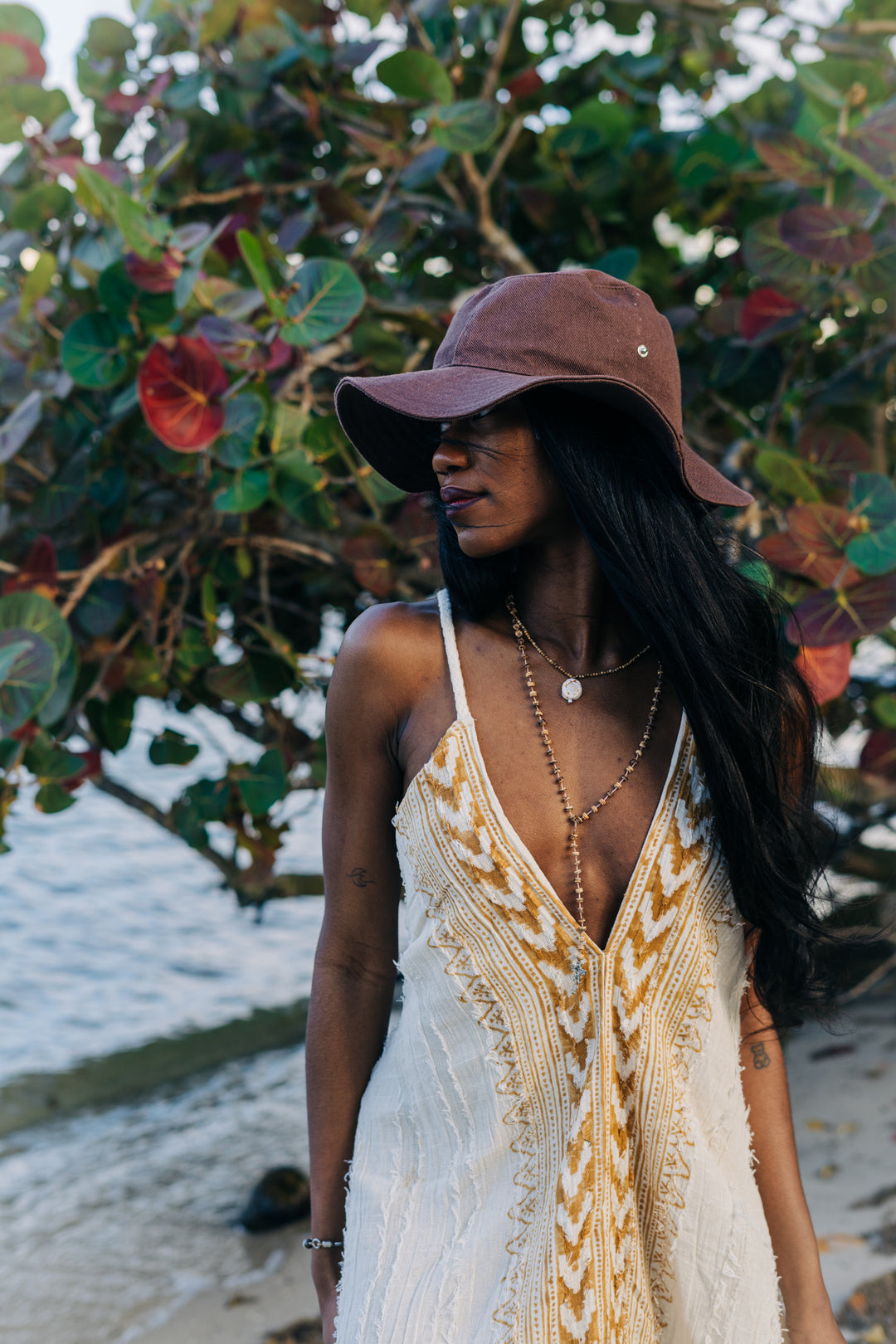 Woman wearing a white and gold dress with a brown hat in front of leafy greenery.