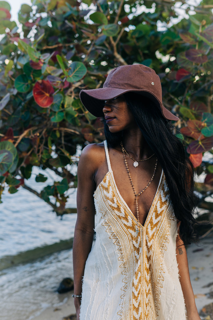 Woman wearing a white and gold dress with a brown hat in front of leafy greenery.