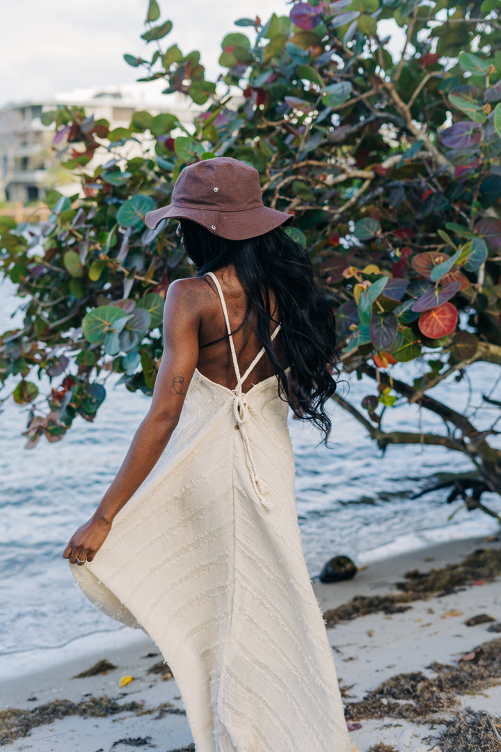 Woman in a beige dress and brown hat walking on a beach with foliage in the background