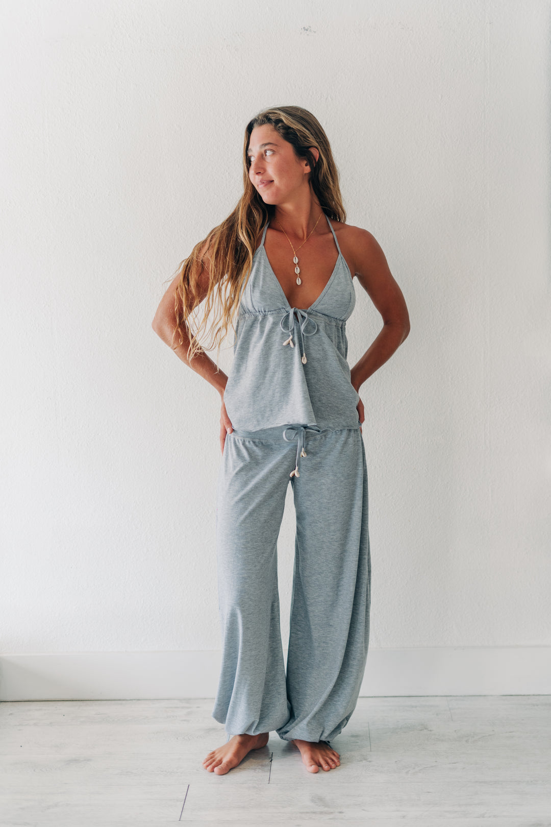A woman modeling a light gray two-piece loungewear set consisting of a halter crop top and matching wide-leg pants, standing against a white wall.