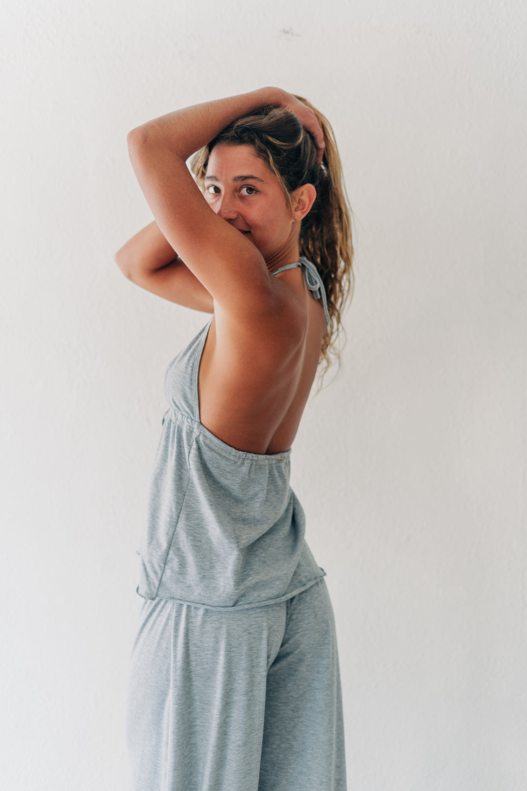 A woman posing in a light gray heathered two-piece loungewear set, featuring a halter-style tank top and matching wide-leg pants, set against a plain white wall.