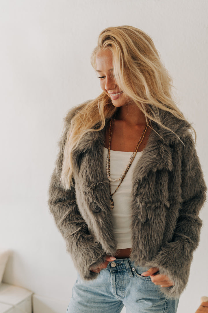 A woman wearing a plush, shaggy faux fur jacket in a medium gray-brown color, styled with a white tank top and light blue jeans against a plain white wall.