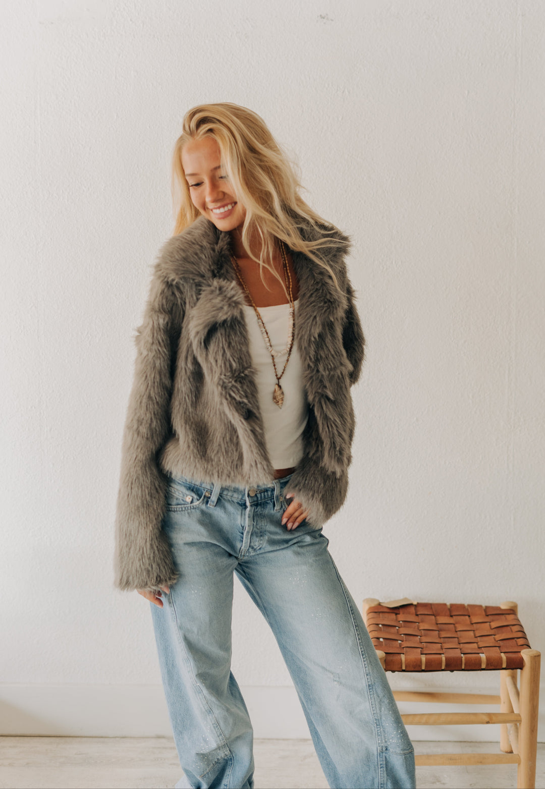 A woman wearing a shaggy gray faux fur jacket, a white tank top, and light blue jeans, standing in a bright room with a woven stool.