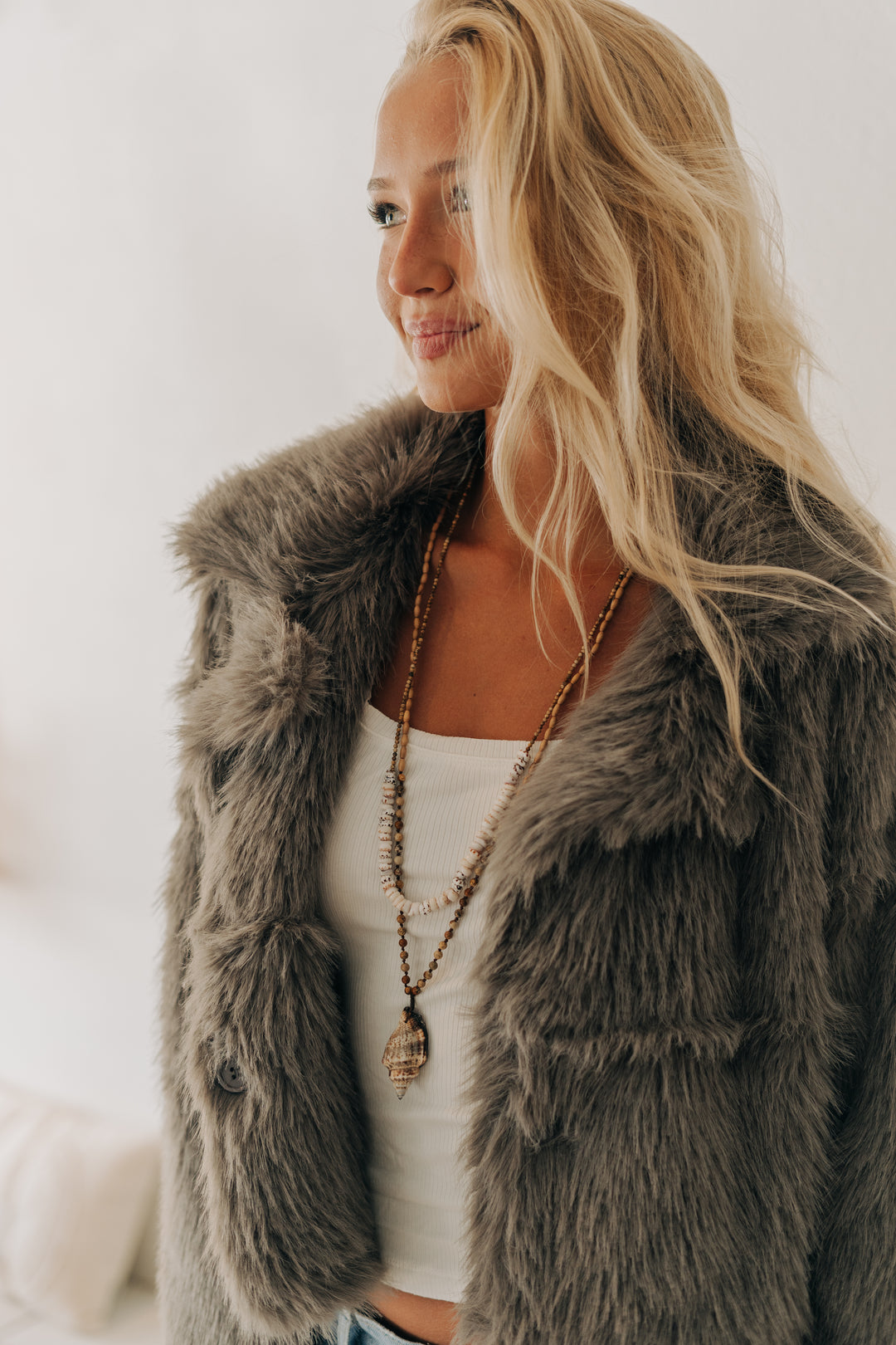 A woman wearing a plush, oversized gray faux fur vest over a white tank top and layered gold necklaces, photographed in a bright, softly lit indoor setting.