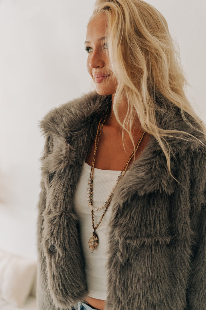 A woman wearing a plush, oversized gray faux fur vest over a white tank top and layered gold necklaces, photographed in a bright, softly lit indoor setting.