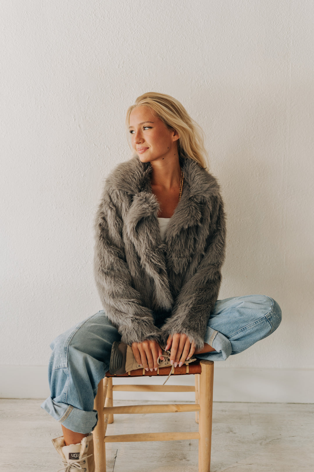 A woman sits cross-legged on a wooden stool wearing a plush gray faux fur jacket, light blue jeans, and white sneakers against a plain white wall.