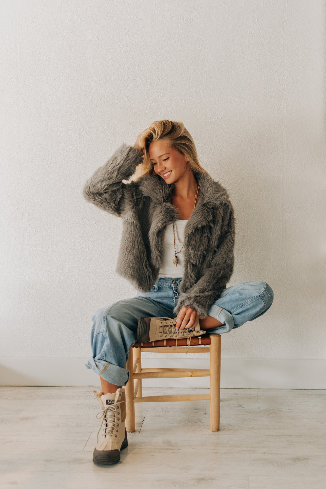 A woman sits cross-legged on a wooden stool wearing a gray faux fur jacket, light blue jeans, and beige lace-up boots against a plain white wall.