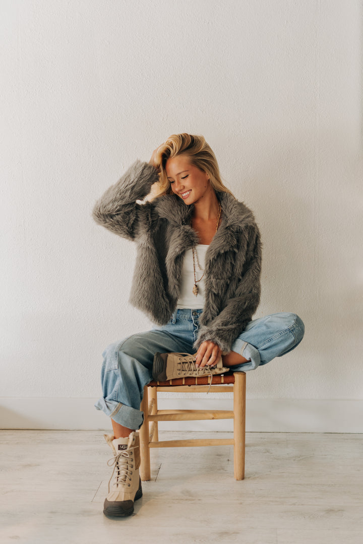 A woman sits cross-legged on a wooden stool wearing a gray faux fur jacket, light blue jeans, and beige lace-up boots against a plain white wall.