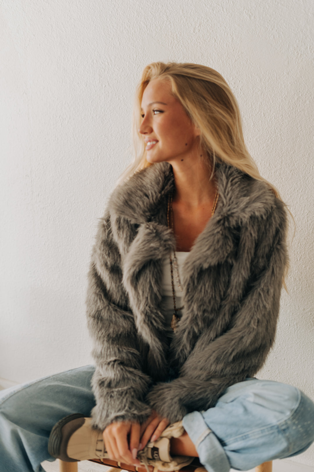 A woman with blonde hair sits wearing a plush gray faux fur jacket, light blue jeans, and brown boots against a textured off-white wall.