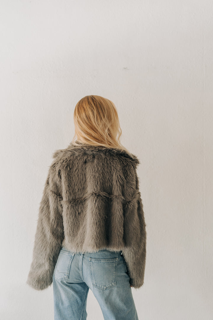 A woman with blonde hair is shown from behind wearing a cropped, textured gray faux fur jacket and light blue jeans, standing against a plain off-white wall.