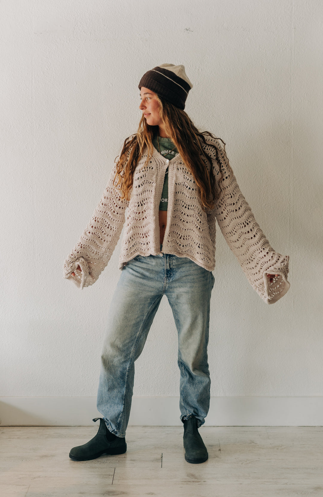 A woman wearing a chunky knit beige cardigan, blue jeans, black boots, and a striped beanie stands in a minimalist white room.