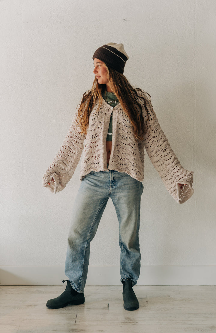 A woman wearing a chunky knit beige cardigan, blue jeans, black boots, and a striped beanie stands in a minimalist white room.