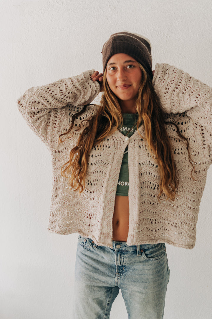 A woman wearing a chunky cream-colored open-front crochet cardigan, a brown beanie, and light-wash blue jeans, posing against a white wall.