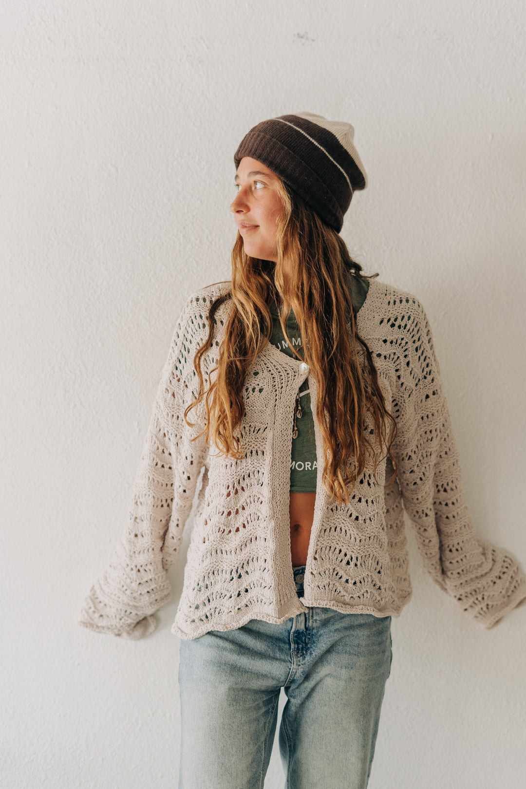 A woman wearing a cream-colored, open-knit crochet cardigan, a dark brown beanie, and light-wash blue jeans, standing against a plain white wall.