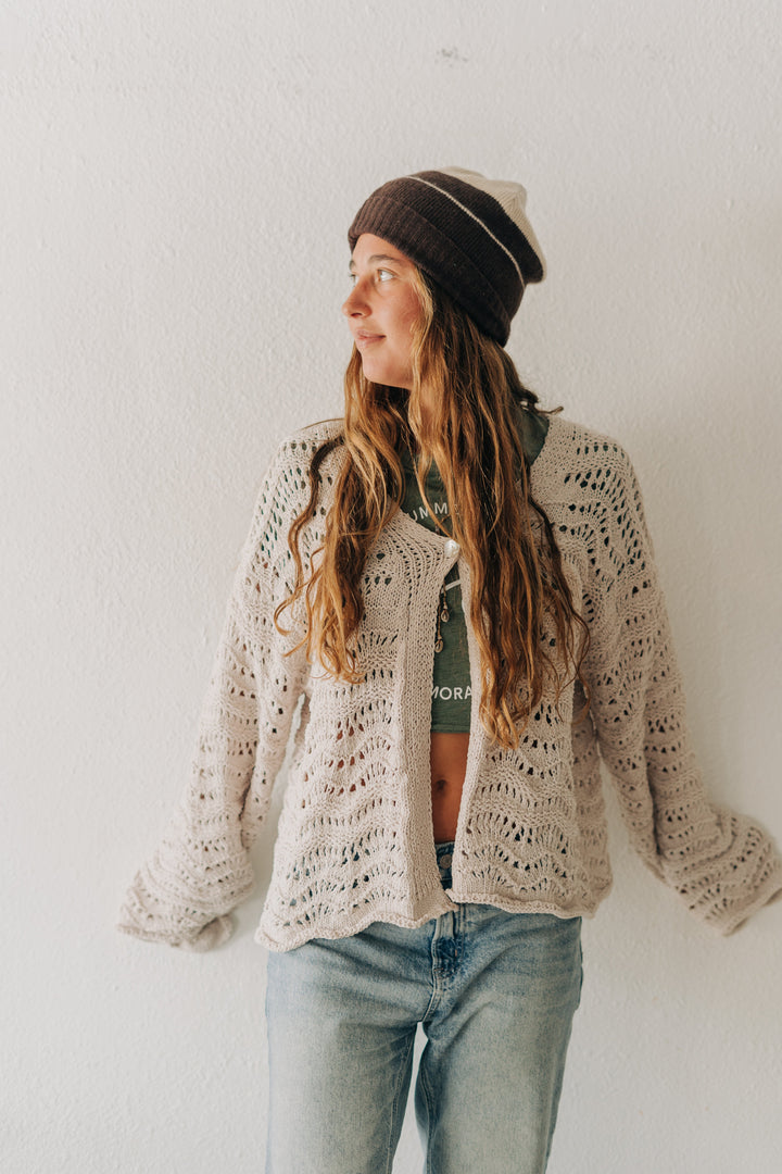 A woman wearing a cream-colored, open-knit crochet cardigan, a dark brown beanie, and light-wash blue jeans, standing against a plain white wall.