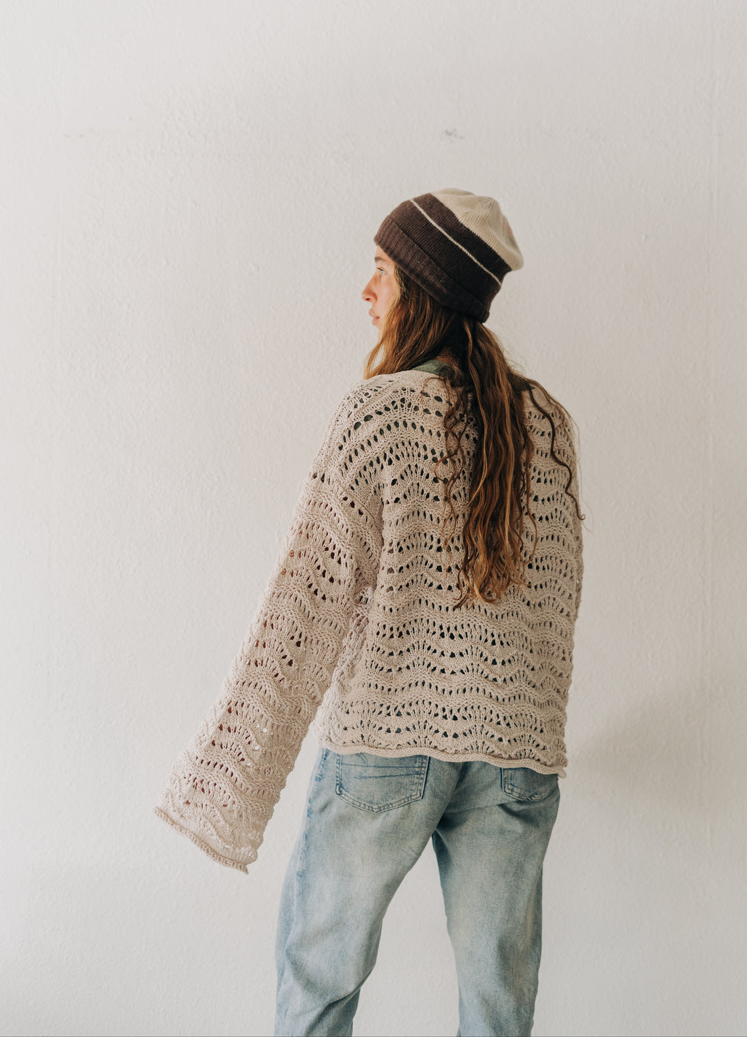 A woman wearing a cream-colored, open-knit crochet sweater, light-wash blue jeans, and a brown and beige striped beanie hat, standing against a plain white wall.